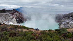 Wide panoramic view of the turquoise acidic crater lake at Mount Ijen in East Java, Indonesia, surrounded by steep rocky cliffs and thick white sulfur smoke, with tourists observing from the rim.