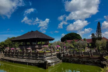 Traditional Bale Kambang floating pavilion surrounded by a calm moat and ornate stone statues at Kerta Gosa Klungkung Palace under a bright blue sky.