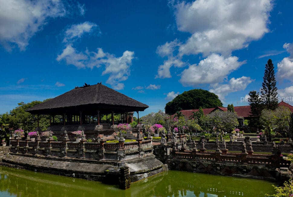 Traditional Bale Kambang floating pavilion surrounded by a calm moat and ornate stone statues at Kerta Gosa Klungkung Palace under a bright blue sky.