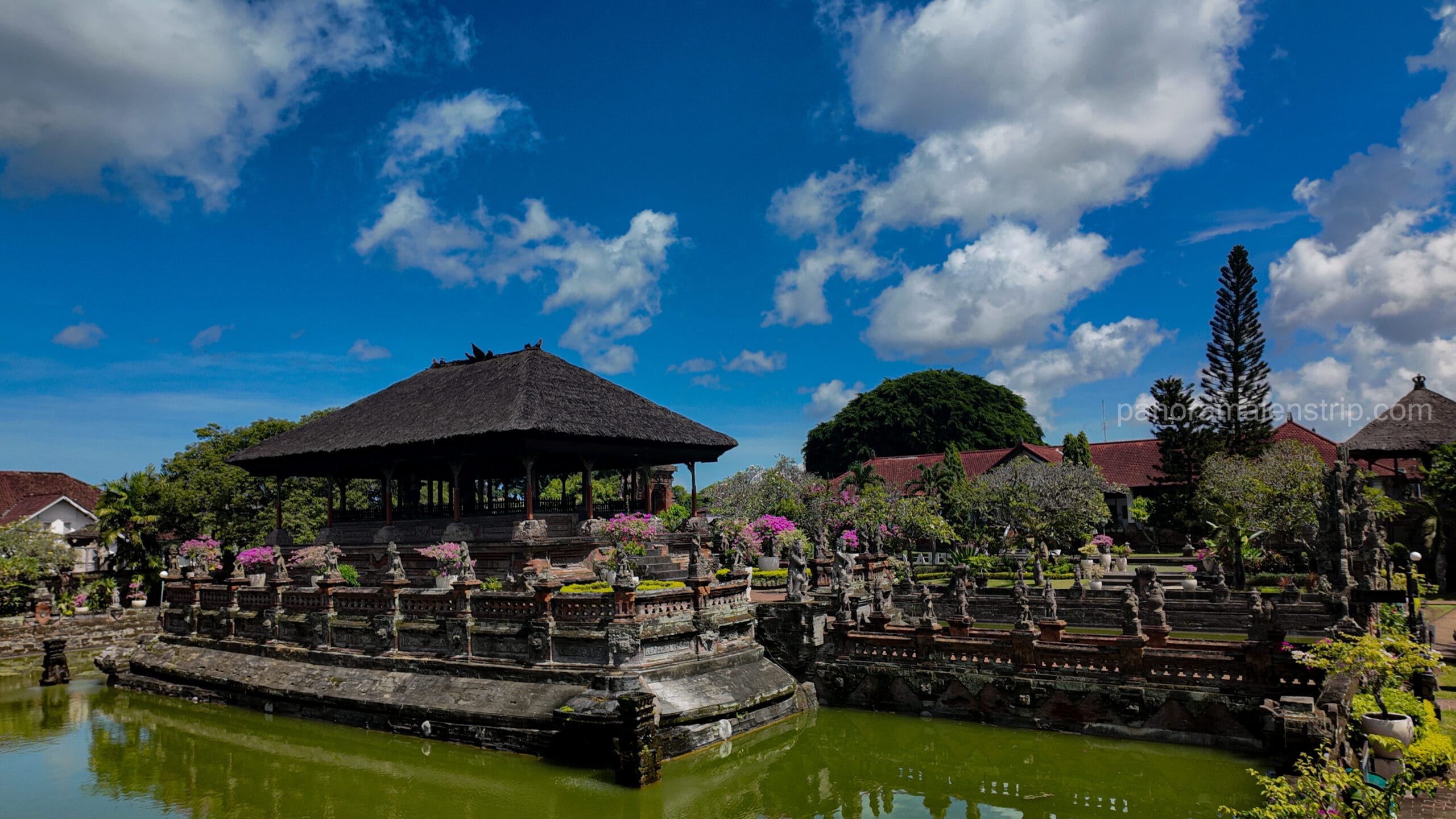 Traditional Bale Kambang floating pavilion surrounded by a calm moat and ornate stone statues at Kerta Gosa Klungkung Palace under a bright blue sky.