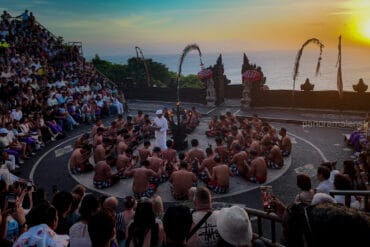 Balinese Kecak fire dance performance at Uluwatu Temple cliff amphitheater during sunset, watched by a large audience of global visitors.