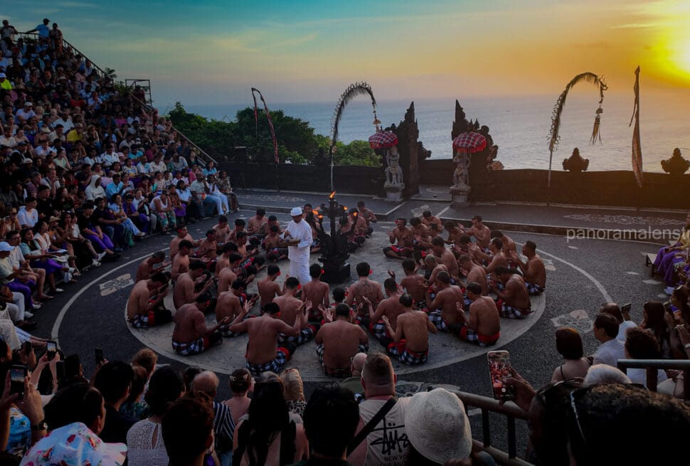 Balinese Kecak fire dance performance at Uluwatu Temple cliff amphitheater during sunset, watched by a large audience of global visitors.