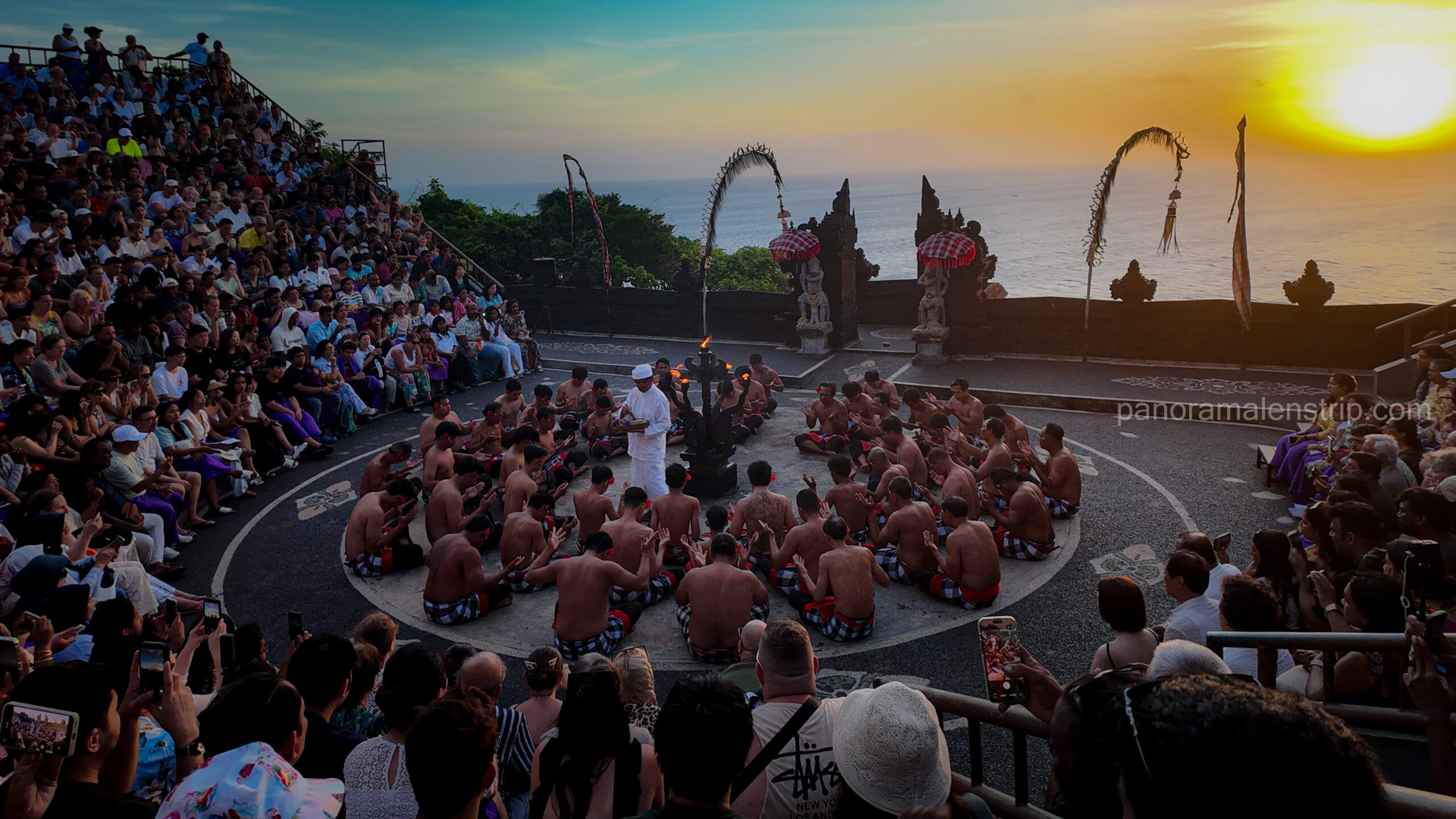 Balinese Kecak fire dance performance at Uluwatu Temple cliff amphitheater during sunset, watched by a large audience of global visitors.