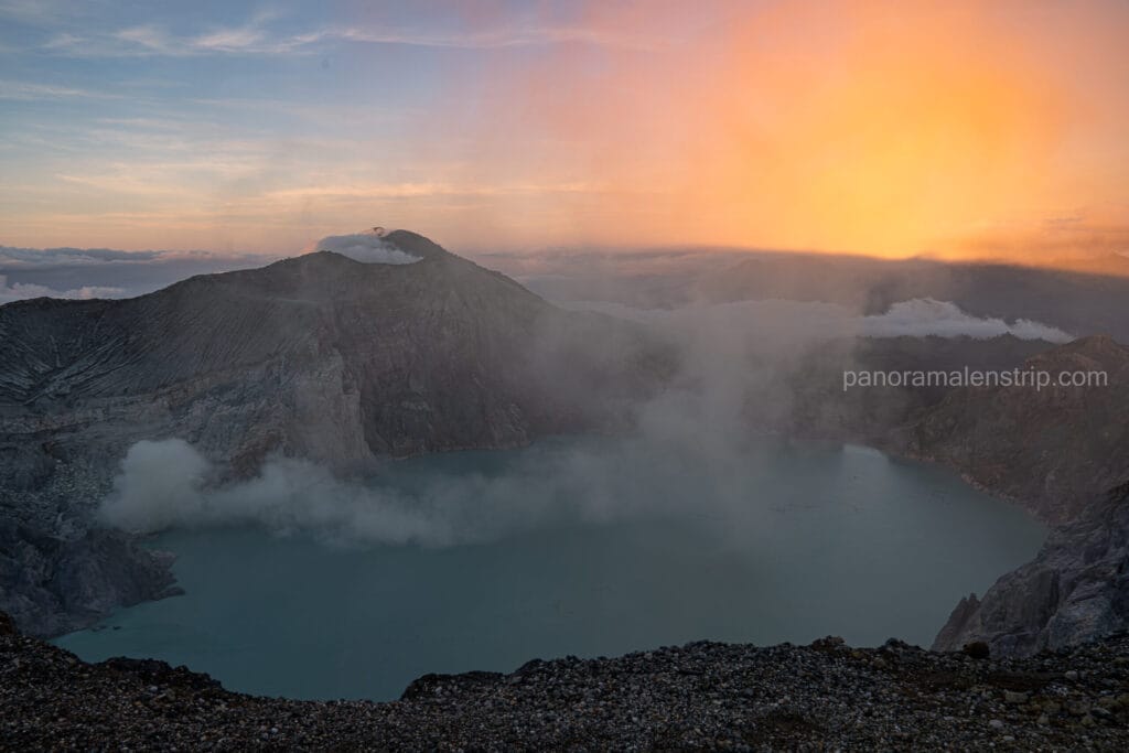 Wide panoramic view of the Kawah Ijen volcanic crater at dawn, showing the vibrant turquoise lake and thick white sulfur smoke rising against a golden sunrise sky in Indonesia.