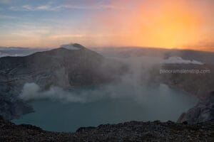 Wide panoramic view of the Kawah Ijen volcanic crater at dawn, showing the vibrant turquoise lake and thick white sulfur smoke rising against a golden sunrise sky in Indonesia.