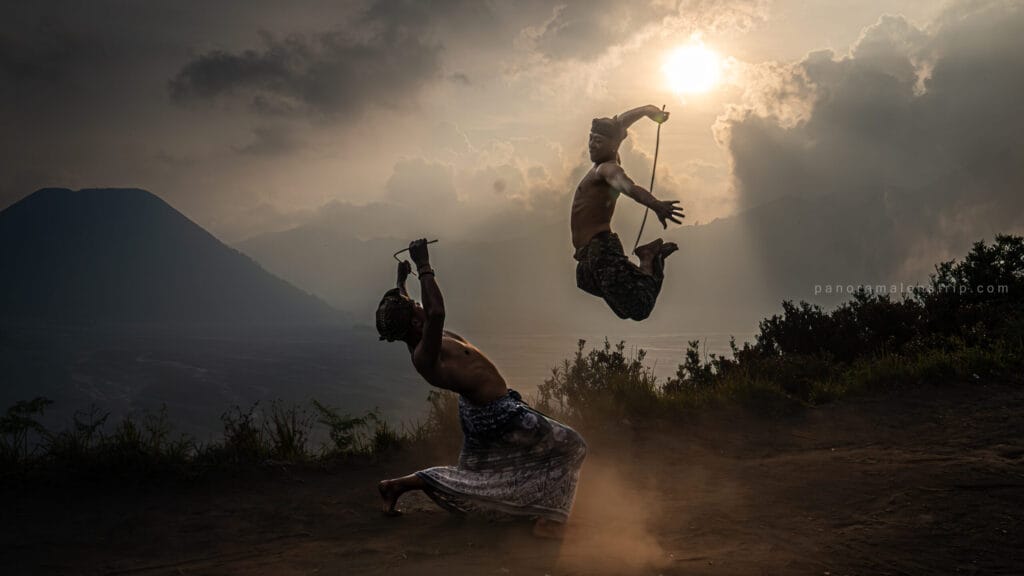 Two shirtless men in traditional sarongs perform a dynamic martial arts sequence with sticks, with one man leaping high into the air against the dramatic, sunlit backdrop of the Mount Bromo caldera.