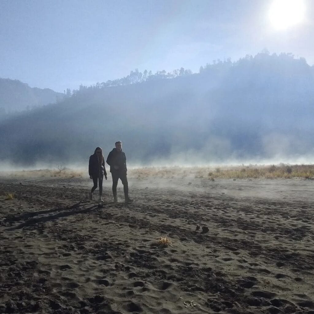 Two hikers walk across misty Bromo volcanic sand sea after sunrise trek to Seruni Point, heading to hotel.