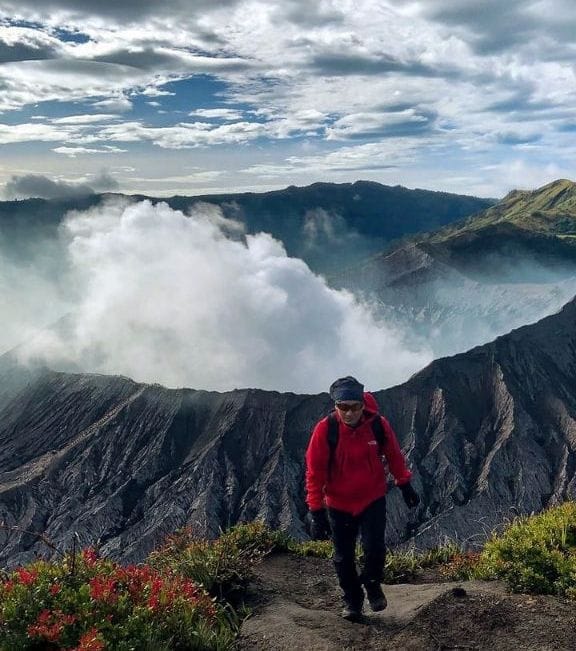 Hiker on Mount Batok trail with Mount Bromo volcano emitting smoke and vast sand sea landscape in Indonesia