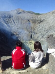 A couple sitting quietly on the edge of Mount Bromo crater, enjoying the smoking volcano view as the culmination of their King Kong Hill trekking journey.