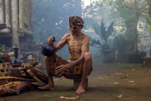 An elderly Balinese man pouring a traditional drink from a clay pitcher into a bamboo cup.