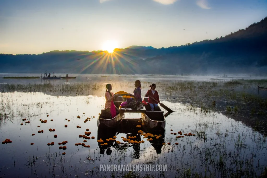 Three Balinese women in a traditional wooden canoe floating on a misty lake during a Bali Culture Tour, surrounded by bright orange marigold flowers.