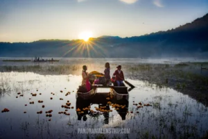 Three Balinese women in a traditional wooden canoe floating on a misty lake at sunrise, surrounded by bright orange marigold flowers.