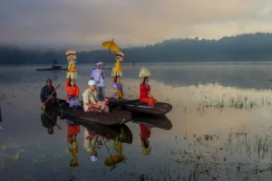Balinese people carrying traditional offerings on a double canoe across a misty lake at dawn.