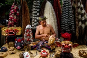 A traditional Balinese artisan carving a wooden mask, surrounded by intricate, colorful ceremonial masks like Barong and Rangda.