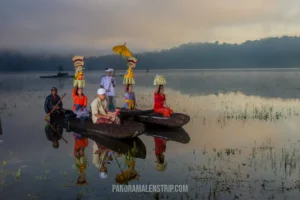 A group of Balinese locals in traditional white and vibrant clothing carrying tall offerings on wooden canoes across a misty lake.