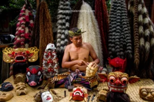 A local craftsman carving a wooden mask while sitting among traditional Barong masks and colorful costume fibers in a Bali artisan village.