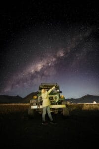 A traveler stands in front of a jeep at Bromo Whispering Sands with the Milky Way and Mount Bromo illuminated under the star-filled night sky.