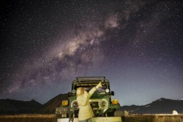 A traveler stands in front of a jeep at Bromo Whispering Sands with the Milky Way and Mount Bromo illuminated under the star-filled night sky.