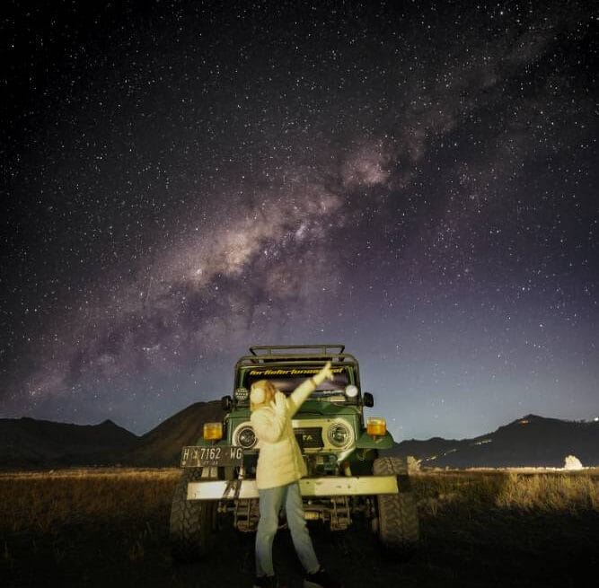 A traveler stands in front of a jeep at Bromo Whispering Sands with the Milky Way and Mount Bromo illuminated under the star-filled night sky.