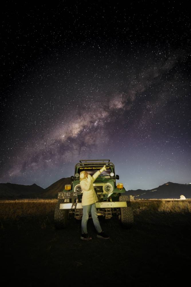 A traveler stands in front of a jeep at Bromo Whispering Sands with the Milky Way and Mount Bromo illuminated under the star-filled night sky.