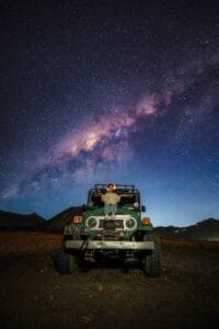 A young child sits on a jeep in the Mount Bromo Sea of Sand parking area, with the Milky Way and Mount Bromo beneath a star-filled night sky.