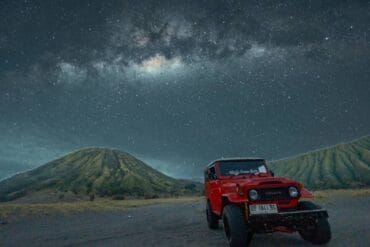 Red jeep parked in front of Mount Batok under a star-filled Milky Way sky at night.