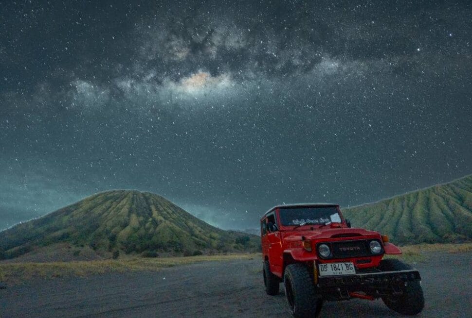 Red jeep parked in front of Mount Batok under a star-filled Milky Way sky at night.