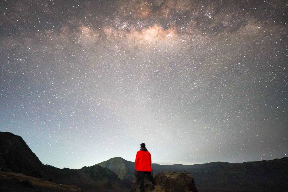 A person in a red jacket standing on the sandy desert of Bromo with the Milky Way glowing overhead at Pasir Berbisik.