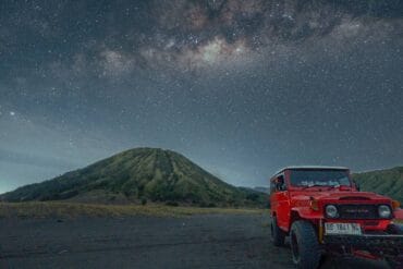Red jeep parked in front of Mount Batok under a starry Milky Way sky at night.