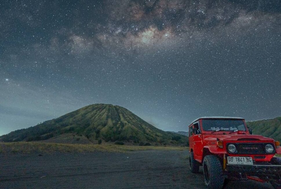 Red jeep parked in front of Mount Batok under a starry Milky Way sky at night.