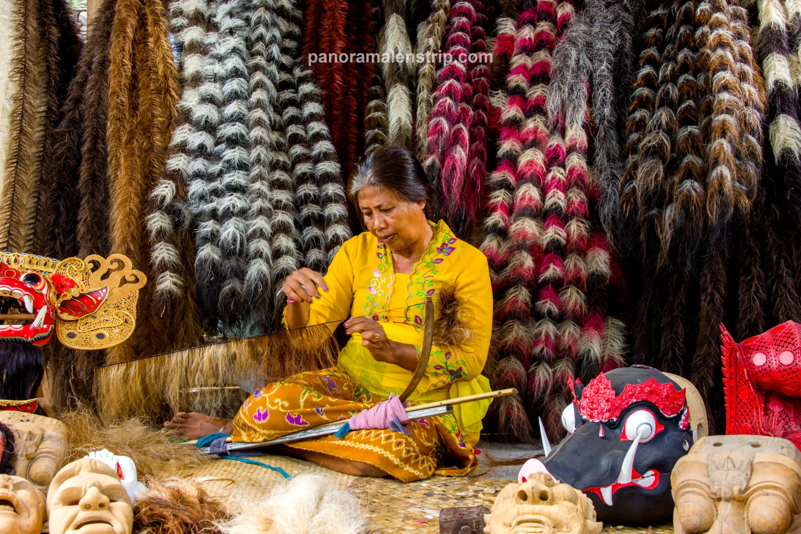 A Balinese woman in a yellow kebaya weaving coarse fibers for a traditional Barong costume inside a vibrant Bali artisan village.