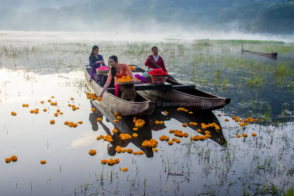 Women in traditional kebayas engaging in a Bali spiritual healing flower offering from a twin-hull canoe on a calm lake.