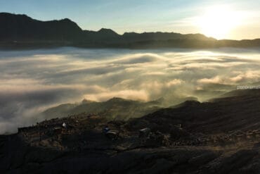A dramatic sunrise over a vast sea of clouds in the Bromo caldera, with silhouettes of tourists and small structures scattered along the dark, rugged volcanic rim in the foreground.