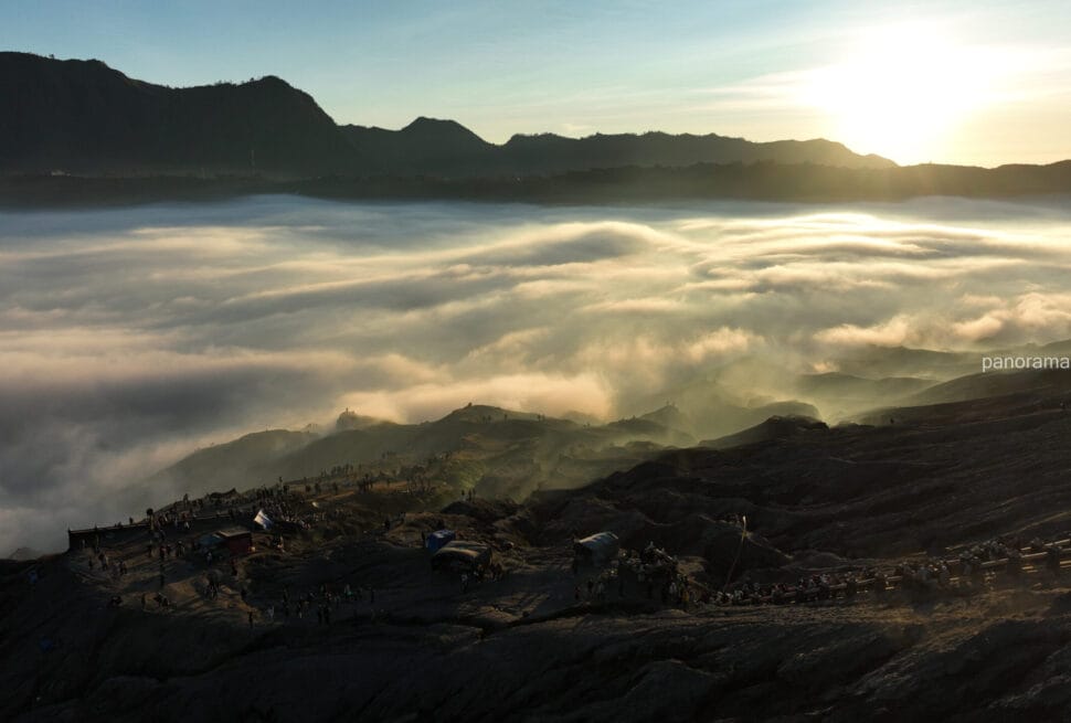 A dramatic sunrise over a vast sea of clouds in the Bromo caldera, with silhouettes of tourists and small structures scattered along the dark, rugged volcanic rim in the foreground.
