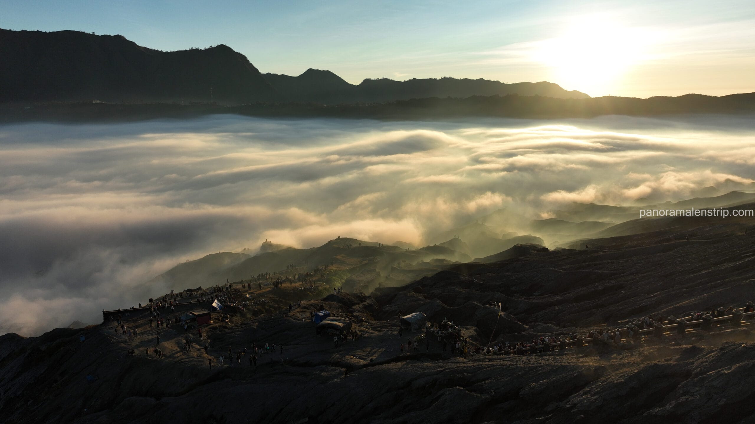A dramatic sunrise over a vast sea of clouds in the Bromo caldera, with silhouettes of tourists and small structures scattered along the dark, rugged volcanic rim in the foreground.