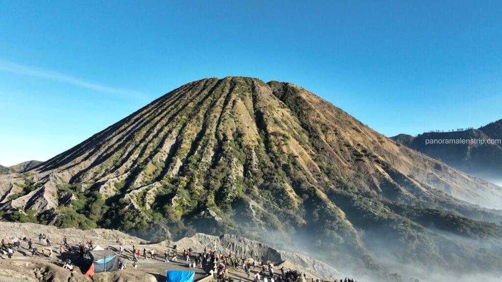 The ribbed volcanic cone of Mount Batok under a clear blue sky, with tourists gathering on the ash terrain at its base in Bromo Tengger Semeru National Park, Indonesia.