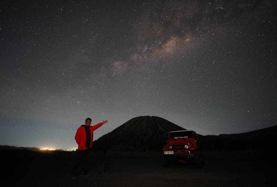 A person in a red jacket stands in front of Mount Widodaren under a starry Milky Way sky at night.