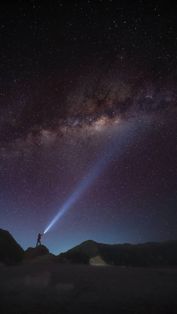A person standing on the sandy dunes of Pasir Berbisik, Bromo, shining a flashlight toward the Milky Way in a star-filled night sky.