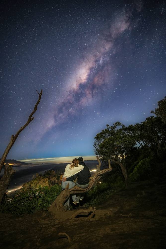 Two people sitting together at Penanjakan Bromo with the Milky Way glowing above Mount Bromo at night.