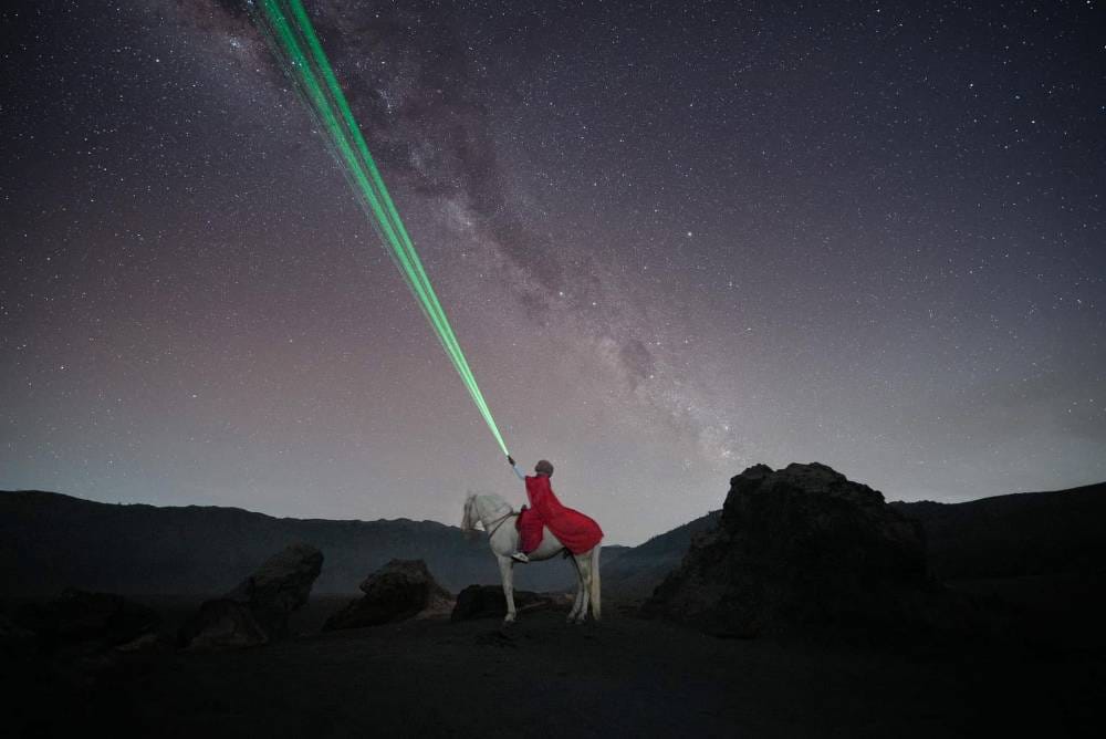 Horse rider in a red cloak on a white horse under the Milky Way at Watu Gede, with green laser beams pointing into the night sky.