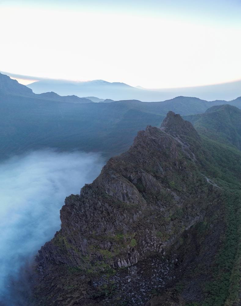 Sunrise at Lembu Suro Cliff Peak in Mount Kelud, Indonesia, with dramatic mountain ridges and low clouds below.