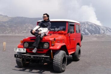 A man sitting on top of a red jeep at Whispering Sands Bromo with Mount Bromo in the background