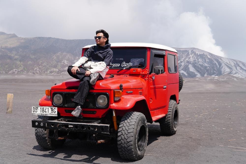 A man sitting on top of a red jeep at Whispering Sands Bromo with Mount Bromo in the background