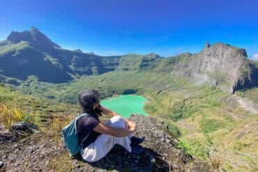 A hiker sits on the summit of Mount Kelud overlooking the crater lake with vivid green water and dramatic volcanic ridges.