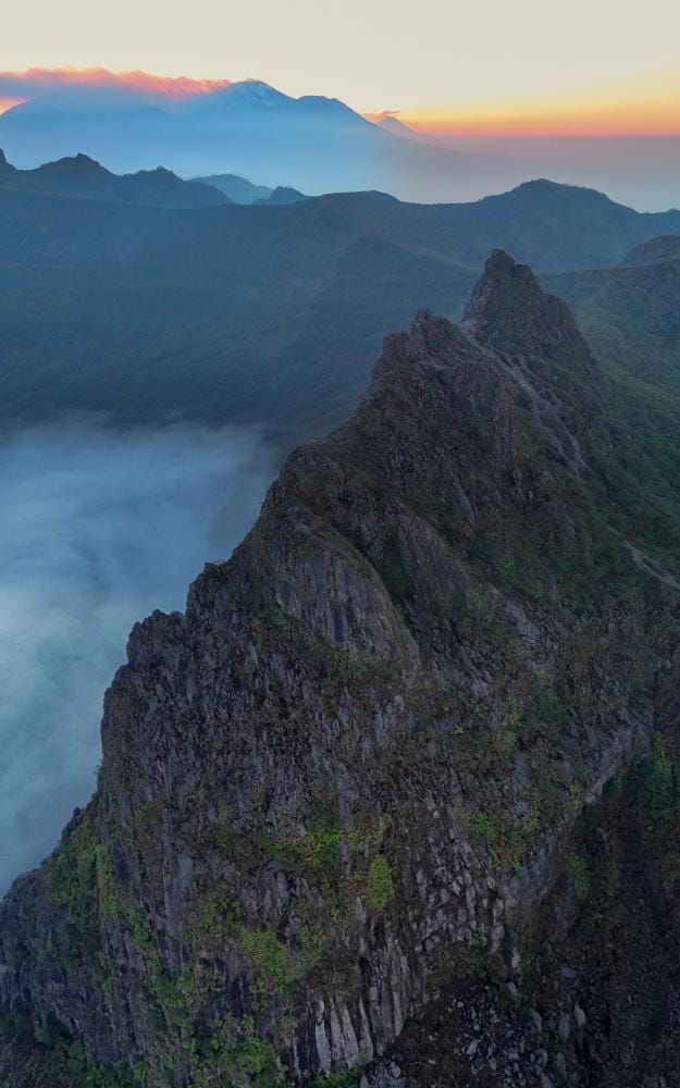 Sunrise over the rugged cliff ridge of Mount Kelud at Puncak Lembu Suro, with mist below and soft dawn light on the horizon.