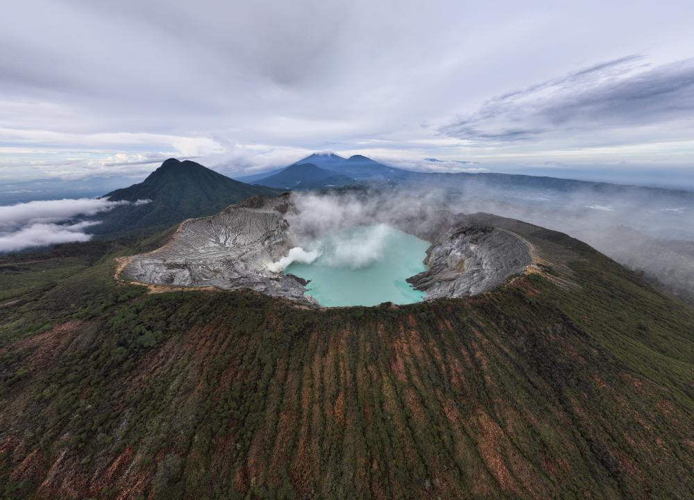 Aerial panoramic view of Kawah Ijen crater lake surrounded by volcanic slopes and drifting mist.