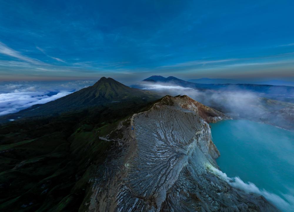 Drone view of sunrise at the entrance to Kawah Ijen, with the crater rim, turquoise lake, and clouds above the volcano