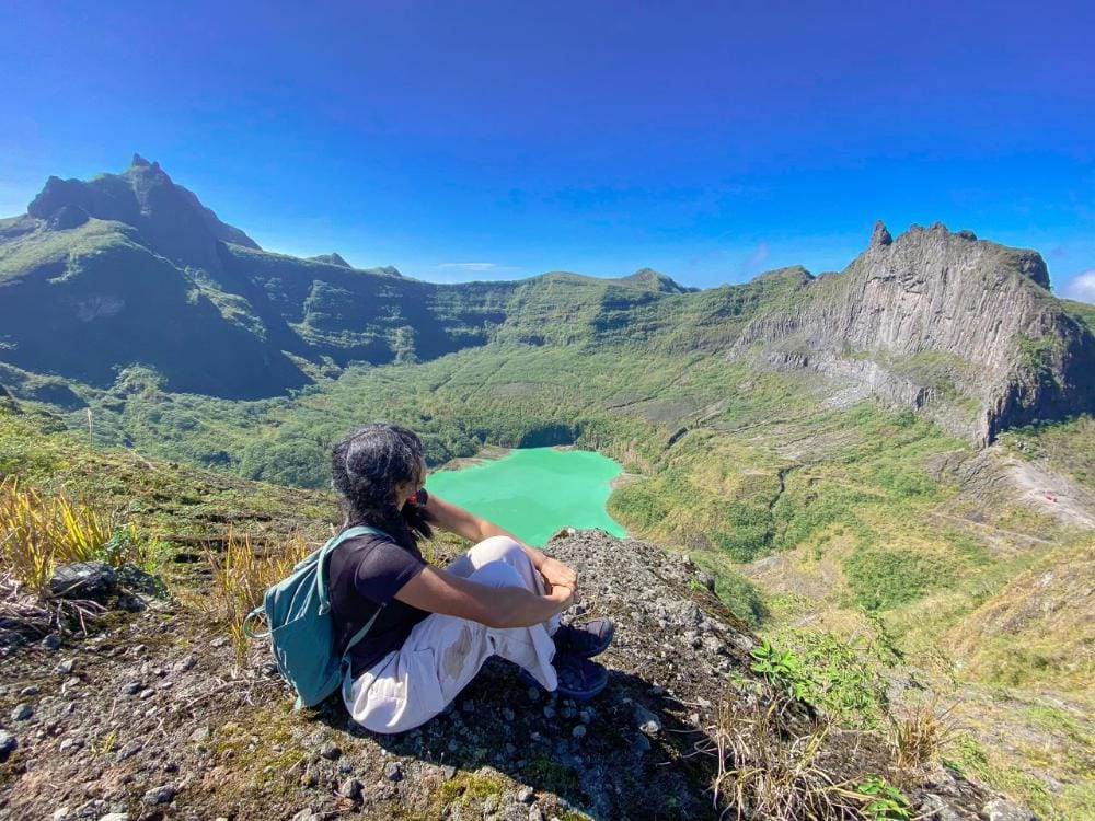 Hiker sitting at the summit of Mount Kelud with a view of the green Kelud Crater Lake in the background